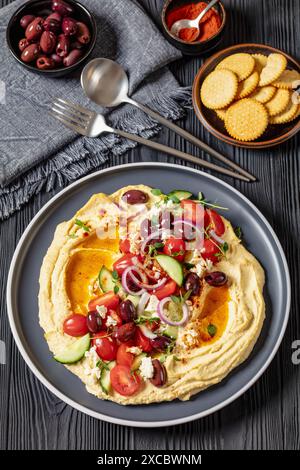 Griechischer Hummus mit frischen Gurken, Oliven, Tomaten, Fetakäse, roten Zwiebeln und Thymian auf Teller auf schwarzem Holztisch, vertikale Ansicht Stockfoto