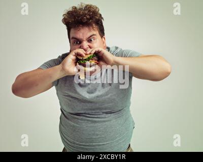 Essen, plus Größe und Mann mit Hamburger im Studio, Hunger und Biss Fast Food mit Mund. Männlicher Mann, Essen zum Mitnehmen und Imbiss zum Verlangen Stockfoto