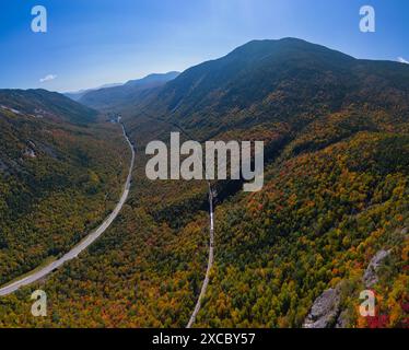 Aus der Vogelperspektive auf ein malerisches Bergtal mit einer gewundenen Straße und lebhaftem Herbstlaub unter einem klaren blauen Himmel. Stockfoto