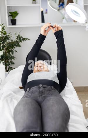 Schöne junge Frau, die ein Selfie mit einem Smartphone macht, während sie sich auf den Tisch legt, direkt nach einer Schönheitsbehandlung und die Ergebnisse genießt Stockfoto