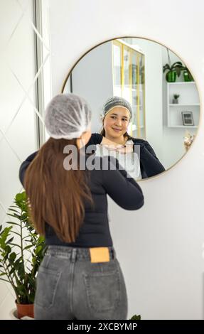 Selbstpflegekonzept. Porträt einer attraktiven jungen Frau, die den Spiegel ansieht, eine schöne Frau, die Eine Einweg-Haarkappe trägt und lächelt und sie genießt Stockfoto