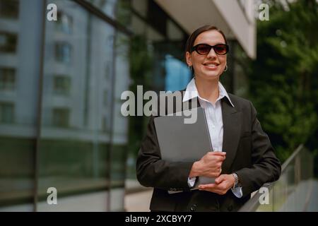 Eine selbstbewusste Geschäftsfrau mit Sonnenbrille hält einen Laptop vor dem Bürogebäude Stockfoto