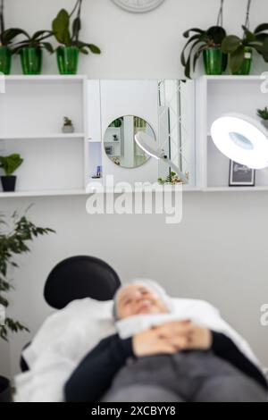 Junge Frau, die sich vor oder nach einem kosmetischen Eingriff auf einem OP-Tisch liegend und lächelte. Die Kreisleuchte beleuchtet den Arbeitsplatz. Stockfoto