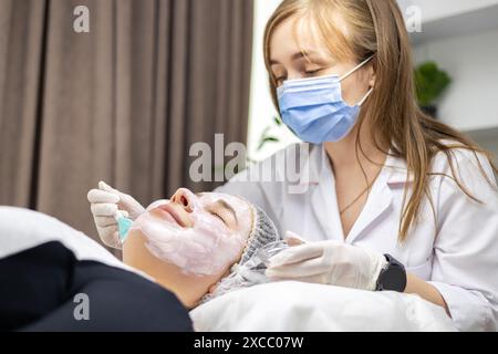 Ein Seitenprofil-Porträt eines jungen Kosmetikers, der weiße Handschuhe und eine chirurgische Maske trägt und eine Gesichtsmaske mit einem Spachtel auf das Gesicht eines Klienten aufträgt. Stockfoto