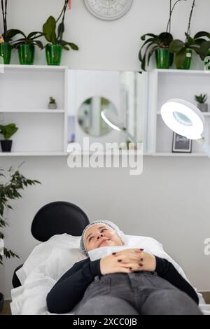 Schöne junge Frau, die sich auf einem OP-Tisch liegend und lächelte, vor oder nach einer kosmetischen Operation. Die Kreisleuchte beleuchtet den Arbeitsplatz. Stockfoto