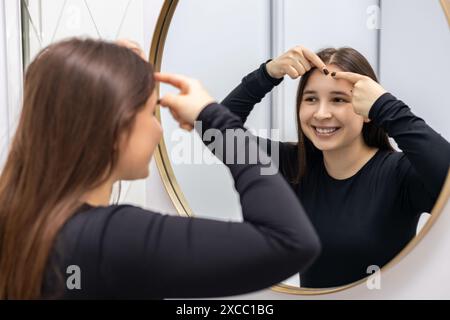 Schöne junge Frau lächelt, während sie im Schrank des Dermatologen einen Pickel auf die Stirn drückt und sich auf eine Schönheitsbehandlung vorbereitet. Stockfoto