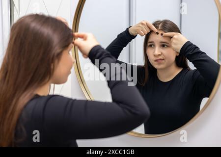 Die junge, schöne Frau im Schrank des Dermatologen schaut sich im Spiegel an und fühlt sich enttäuscht von der Qualität ihrer Haut. Stockfoto