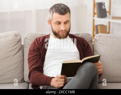 Bärtiger Mann sitzt auf der Couch und liest Buch Stockfoto