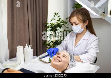 Junge, erfahrene Dermatologin, die die Spritze und Flasche Lösung für die folgende chemische Peeling- und Schönheitsbehandlung vorbereitet, während ihr Kunde ist Stockfoto