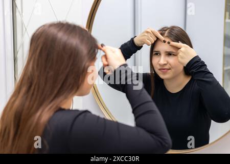 Eine junge Frau in einer schwarzen Bluse, die unzufrieden aussieht, untersucht ihre Haut und analysiert ihren Zustand vor einer Schönheitsbehandlung, während sie in die schaut Stockfoto