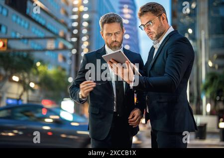 Geschäftsleute prüfen ihre E-Mails und lesen wichtige Dokumente auf einem Tablet vor dem Hintergrund einer Stadt bei Nacht. Zwei Geschäftsleute benutzen Tablets, um im Freien zu arbeiten Stockfoto