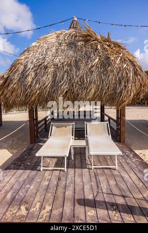 Nahaufnahme einer mit Palmblättern bedeckten Cabana am Sandstrand eines Hotels, ausgestattet mit einem Bett, Liegestühlen und einem Getränketisch. Curacao. Stockfoto