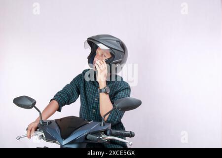 Reifer asiatischer Mann posiert auf dem Motorrad, spricht während der Pause auf dem Handy, trägt Helm, schaut seitwärts mit verwirrtem Ausdruck, isoliert auf weiß Stockfoto