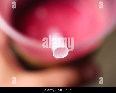 Ein Makrofoto von der Spitze eines Strohs, der in eine Tasse mit rosafarbenem Getränk eingelegt wurde. Der Becher wird in einer Hand auf dem unscharfen Hintergrund gehalten. Stockfoto