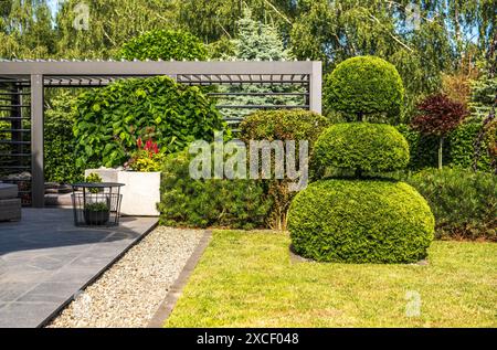 Blick auf einen gepflegten Hinterhof mit einer Pergola und üppigem Grün. In der Mitte steht ein großer, getrimmter Busch, der wie ein Schneemann geformt ist Stockfoto