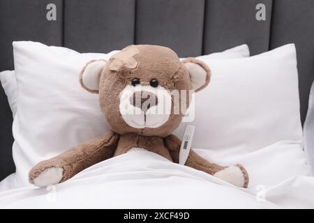 Niedlicher Bär mit klebendem Pflaster und Thermometer unter der Decke im Bett Stockfoto