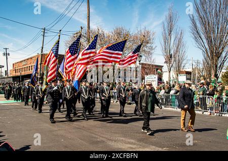 Lindenhurst, New York, USA, 30. März 2024 - uniformierte Marschierer mit American Flags gehen stolz auf Eine Sunny Street während Einer Parade, an der sich Eine lebhafte Menge von Zuschauern an den Seiten aufeinanderreiht, um Ein patriotisches Ereignis in einem urbanen Ambiente zu feiern. Stockfoto