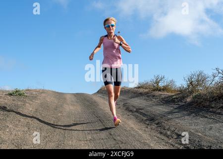 Sportliche Frau in rosa gekleidet, die auf einer abgelegenen Wüstenstraße bergab läuft, mit blauem Himmel im Hintergrund. Stockfoto