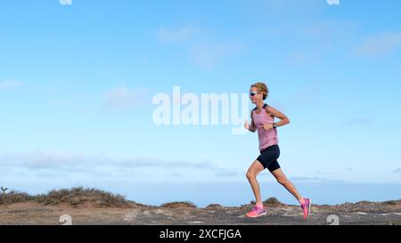 Eine Frau in einem pinkfarbenen Oberteil und schwarzen Shorts rennt durch eine einsame Landschaft und hält auf felsigen Pfaden ein ruhiges Tempo. Ihr lebhaftes Outfit steht im Kontrast Stockfoto