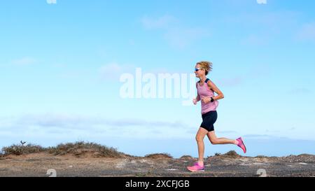 Eine Frau in einem pinkfarbenen Oberteil und schwarzen Shorts rennt durch eine einsame Landschaft und hält auf felsigen Pfaden ein ruhiges Tempo. Ihr lebhaftes Outfit steht im Kontrast Stockfoto