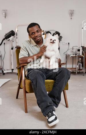Vertikales Porträt des jungen Schwarzen Mannes, der den niedlichen Spitz-Hund im Schoß hält, während er im Pflegestudio auf dem Stuhl sitzt Stockfoto