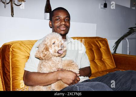 Minimales Porträt eines afroamerikanischen Mannes mit niedlichem Hund auf dem Schoß, der auf der Couch sitzt und in die Kamera blickt, mit Blitz Stockfoto