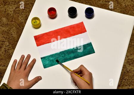 Ein Junge zieht die Flagge Ungarns auf ein weißes Blatt mit Pinsel und Farben. Stockfoto