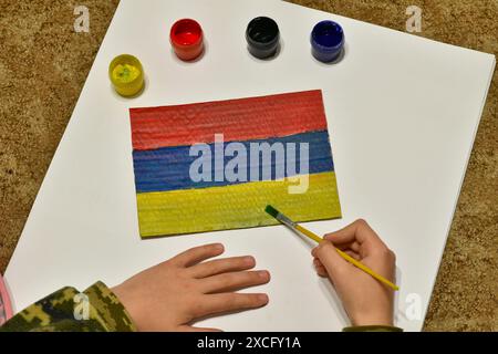 Der Junge auf dem Boden zieht die Flagge Armeniens mit einem Pinsel und malt auf Papier. Stockfoto