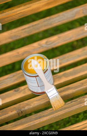 Pinsel und Dose Farbe auf einer Holzbank im Freien öffnen. Die Farbe ist goldgelb und die Bank ist aus natürlichem Holz. Stockfoto