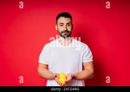 Porträt eines jungen bärtigen Mannes, der Zitronen in beiden Händen auf einem isolierten roten Hintergrund hält Stockfoto