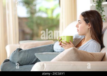 Nachdenkliche Frau, die zu Hause Kaffee trinkt und durch ein Fenster in den Garten blickt Stockfoto