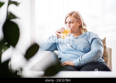 Porträt einer schönen jungen übergewichtigen Frau zu Hause, die Orangensaft trinkt, süßen Geschmack genießt. Schönheits- und Körperpositivitätskonzept. Stockfoto