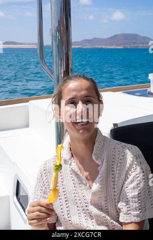 Ein junger Mann lächelt hell, während er einen Snack auf einem Boot genießt, das weite blaue Meer und die ferne Küste bieten eine malerische Kulisse. Stockfoto