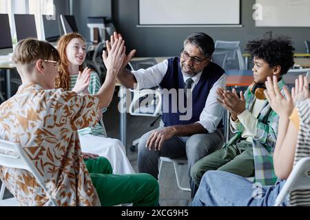 Fröhlicher, reifer Schulpsychologe, der High-Five mit männlichen Schülern während der Gruppentherapie macht, andere Teenager klatschen die Hände Stockfoto