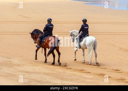 Gijon, Spanien - 24. Mai 2024: Zwei bewaffnete spanische Polizisten in ordentlichen Uniformen reiten auf wunderschönen Pferden an einem hellen sonnigen Tag patrouillieren an einem Sommerstrand Stockfoto