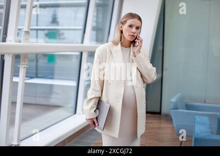 Porträt einer schönen schwangeren Geschäftsfrau in einem weißen Anzug, die in einem hellen Büro auf einem Handy mit einem Laptop vor einem weiß-blauen Ba spricht Stockfoto
