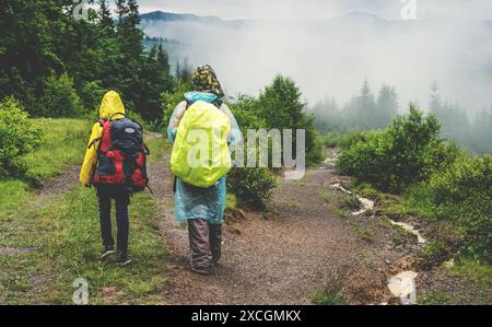 Zwei Wanderer im Regenmantel wandern auf dem Weg zum grünen Bergwald im Nebel mit dem gelben Rucksack bei regnerischem Wetter. Stockfoto