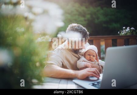 Papa arbeitet im Homeoffice, sitzt auf der Terrasse mit Laptop und hält das kleine Mädchen. Vater verbringt Zeit mit einer kleinen Tochter und genießt die gemeinsame Zeit Stockfoto