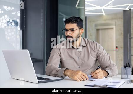 Seriöser und konzentrierter Geschäftsmann, der Webinar-Onlinekurs ansieht, Mann, der im Büro am Arbeitsplatz aufrüstet, hispanischer Mann, der Notizen macht und Daten in Notizbücher schreibt Stockfoto