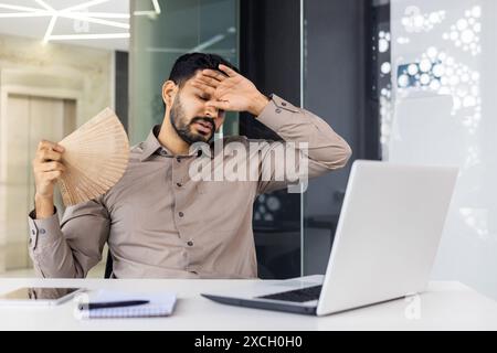 Geschäftsmann in braunem Hemd fächert sich an, fühlt sich aufgrund der Hitze in einem modernen Büro gestresst, während er an einem Laptop arbeitet. Stockfoto