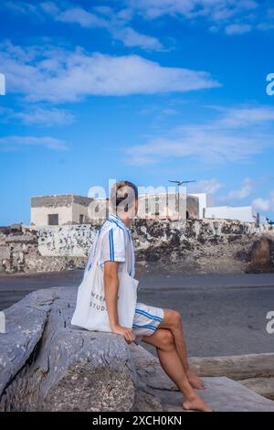 Ein junger Mensch sitzt auf einem Treibholz und blickt auf das Meer mit blauem Himmel und weißen Wolken darüber. Stockfoto