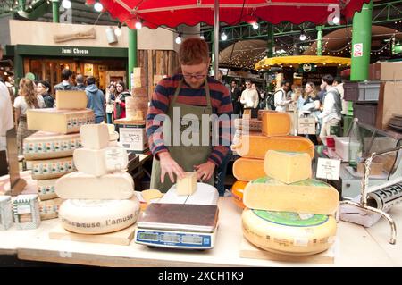 Käsestand, Borough Market, Bermondsey, Southwark, London, Großbritannien - französische und niederländische Käsesorten - Gouda; Mann, der Käseportionen schneidet; viele Menschen im Hinterkopf Stockfoto