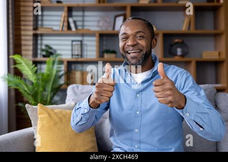 Fröhlicher Mann, der lächelt und die Daumen nach oben drückt, während er auf einem Sofa sitzt, in einer gemütlichen Wohnumgebung, mit Regalen und Pflanzen im Hintergrund. Stockfoto