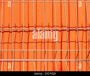 Abstrakt mit Muster eines profilierten Metallgitters, der einen Schatten auf eine orange lackierte profilierte Metallschutzwand wirft. Hintergrund. Stockfoto