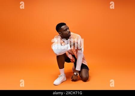 Hübscher afroamerikanischer Mann in modischer Kleidung, der sich auf lebhaftem orangefarbenen Hintergrund hockt. Stockfoto