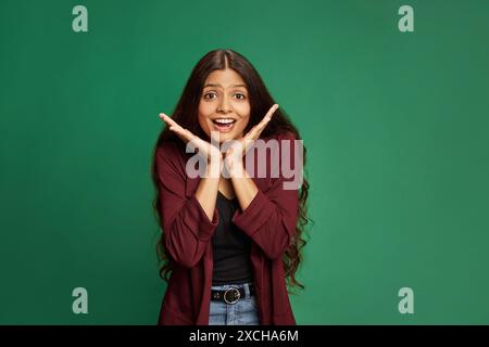 Porträt einer überraschten und staunenden jungen Indianerin, die mit offenem Mund in die Kamera schaut und Hände vor grünem Studiohintergrund spreizt. Stockfoto