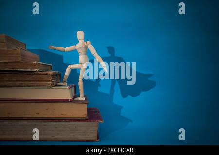 Hölzerne Schaufensterpuppe klettert Treppen aus Vintage-Büchern und projiziert einen Superhelden-Schatten mit einem cape auf einem blauen Hintergrund Stockfoto