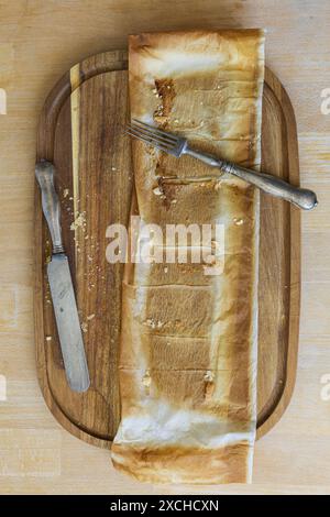 Tischfotografie eines Schneidebretts aus Akazienholz mit Backpapier, das vom Kuchen übrig geblieben ist, und Vintage-Gabel und Messer auf einem Holztisch Stockfoto