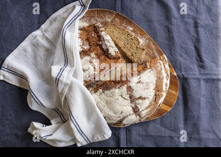 Tischfotografie eines frisch gebackenen Roggenbrotes, das auf einem Schneidebrett aus Akazienholz in zwei Hälften geschnitten und in Scheiben geschnitten wird, bedeckt mit einem weißen Küchentuch auf einem dunklen B Stockfoto