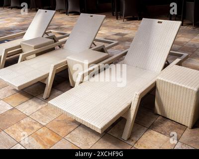 Drei leere, moderne Sonnenliegen aus weißem Rattan, Liegestühle mit Beistelltisch für Gäste auf gelbem Fliesenboden am Außenpool im Hotelresort mit Stockfoto